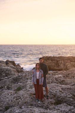 Çift deniz kenarındaki taşların üzerinde duruyor, sarılıyor, manzaranın ve temiz havanın tadını çıkarıyor..