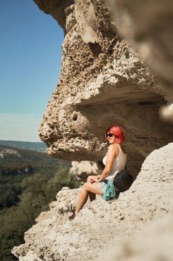 Genç bir kadın bir kayanın üzerinde oturur, manzaraya bakar ve manzaranın ve temiz havanın tadını çıkarır..