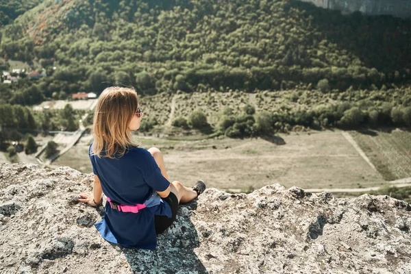 Jovem mulher senta-se em uma rocha, olhando para a paisagem e apreciando a vista e ar fresco. — Fotografia de Stock