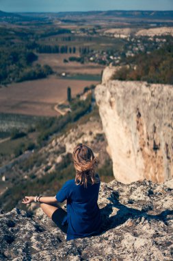 Genç bir kadın bir kayanın üzerinde oturur, manzaraya bakar ve manzaranın ve temiz havanın tadını çıkarır..