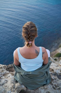 Kadın bir kayanın üzerinde oturuyor, deniz manzarasına, gün batımına bakıyor ve manzaranın ve temiz havanın tadını çıkarıyor..