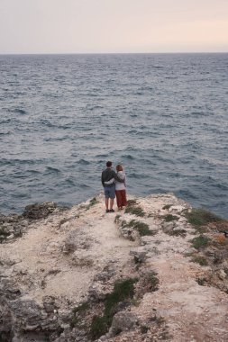 Çift denizin kenarındaki bir kayanın üzerinde duruyor, sarılıyor, manzaranın ve temiz havanın tadını çıkarıyor..