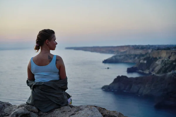 Mujer se encuentra en una roca, mirando el paisaje marino, puesta de sol y disfrutando de la vista y el aire fresco. — Foto de Stock