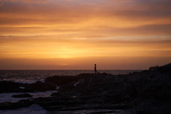 Mulher está de pé sobre as rochas junto ao mar, no spray do surf, desfrutando do pôr do sol e do ar fresco. — Fotografia de Stock