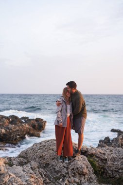 Çift deniz kenarındaki taşların üzerinde duruyor, sarılıyor, manzaranın ve temiz havanın tadını çıkarıyor..