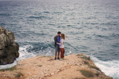 Çift denizin kenarındaki bir kayanın üzerinde duruyor, sarılıyor, manzaranın ve temiz havanın tadını çıkarıyor..