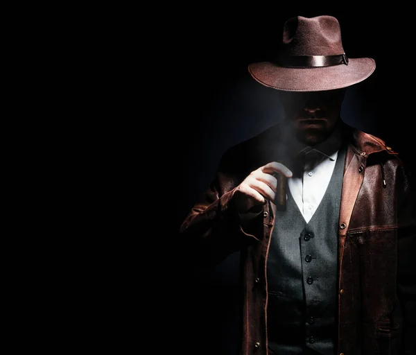 stock image Photo of a shaded detective in jacket and hat holding and smoking cigar on black background.