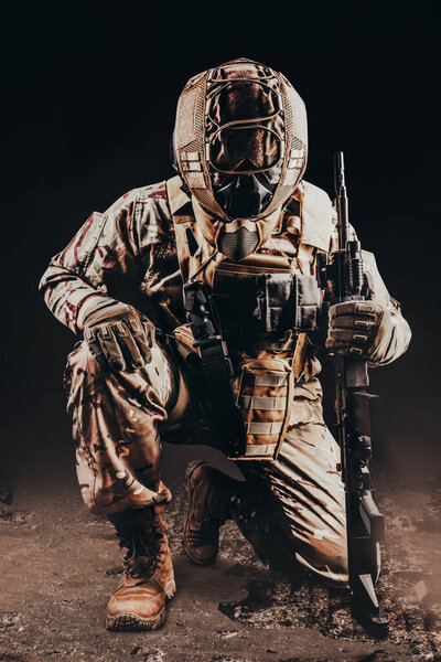 Photo of a fully equipped soldier in armored vest. helmet and camouflaged outfit kneeling with rifle on black background.