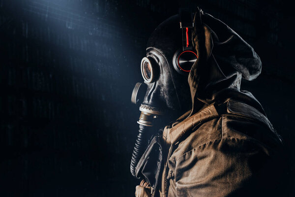 Photo of a stalker soldier in soviet rubber gas mask with hose, wearing headphones and standing profile view on dark underground backdrop.