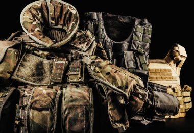 Various types of soldier camouflaged armored vests with shoulder and neck protection on black background laying on table. clipart