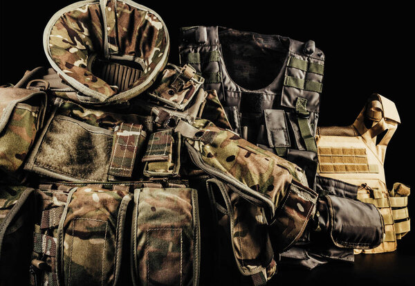Various types of soldier camouflaged armored vests with shoulder and neck protection on black background laying on table.