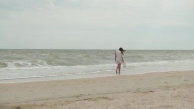 Kız sahilde deniz kabuğu topluyor. Sahilde yürüyen biri.