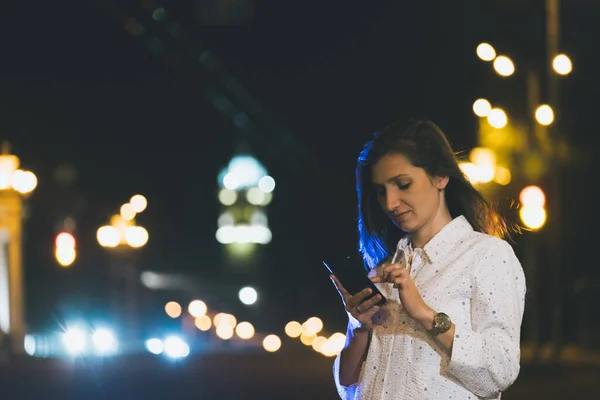 Chica atractiva en traje blanco usando teléfono inteligente en la noche,, mujer joven sonriendo con el móvil en las manos —  Fotos de Stock