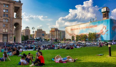 Görüntü canlı bir şehir manzarası gösteriyor. İnsanların dinlendiği ve oturduğu çimenli bir alan var. Bazıları çimlerin üzerinde güneşli bir günün tadını çıkarıyor. Bu sakin kalabalığın arkasında, yoğun bir kalabalık var.