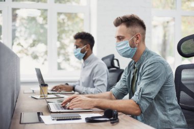 Ofiste çalışan bir grup genç iş adamı tıbbi koruyucu maskeler takıyor. İş arkadaşları birlikte çalışırken dizüstü bilgisayar kullanırlar. Kişisel bakım, koronavirüs COVID-19 koruma, sosyal uzaklık kavramı