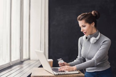 Kredi kartı tutan ve dizüstü bilgisayar kullanan genç bir kadın. İnternette çalışan bir kız. Çevrimiçi alışveriş, e-ticaret, internet bankacılığı, harcama, ev konseptinden çalışma