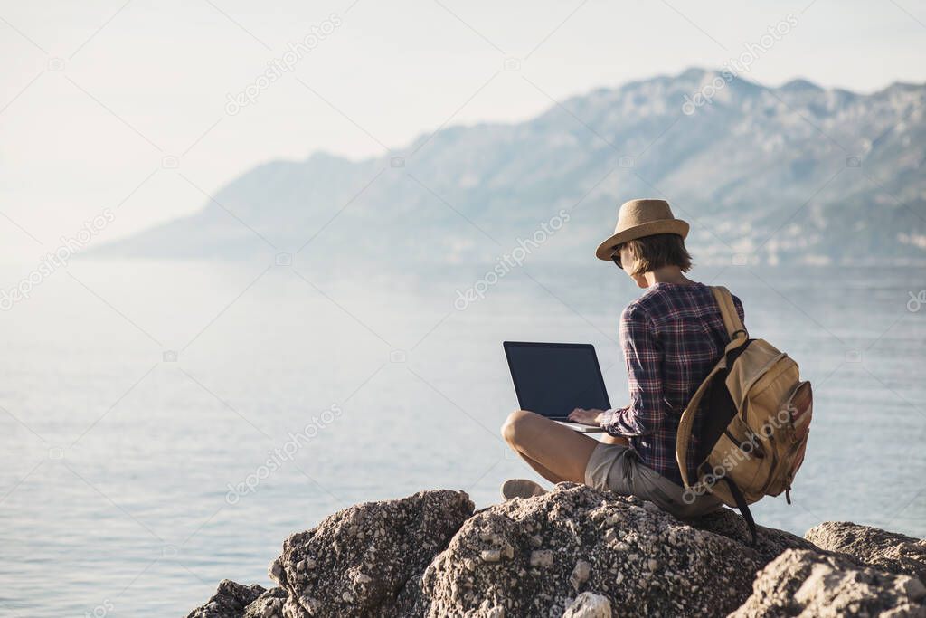 Young woman using laptop computer by the sea. Freelance work concept. People using devices to plan trips, check in to hotels and flights, stay connected with family and office from remote part of the world