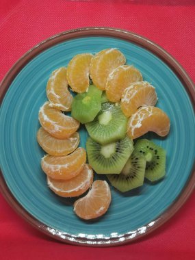 kivi, muz, mandalina, yuvarlak turkuaz bir tabakta, kırmızı arka planda kahverengi bir kenarı var.