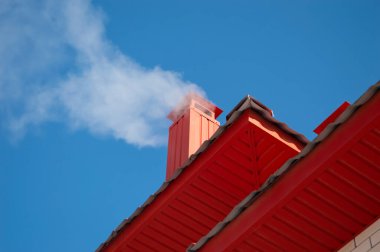 Yakın plan - kırmızı çatıdaki bacadan gelen duman soğuk bir günde, mavi gökyüzüne karşı