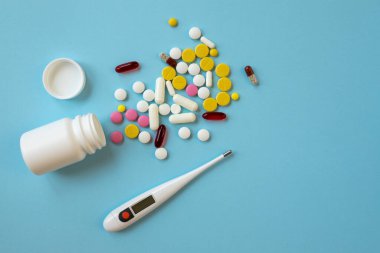 Mavi arka planda çok renkli tabletler, haplar, kapsüller ve dijital termometre. Hasta bir insan için tedavi kavramı. Üst görünüm