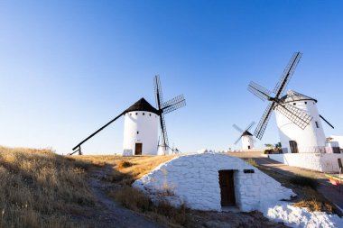 Tarihsel yel değirmenleri ve bir mağara evi açık mavi gökyüzünün altındaki kuru manzarayı noktalıyor.