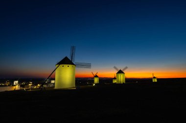 Geleneksel İspanyol yel değirmenleri alacakaranlıkta aydınlanan bir tepenin üzerinde canlı bir günbatımına ve mavi gökyüzüne karşı duruyorlar.