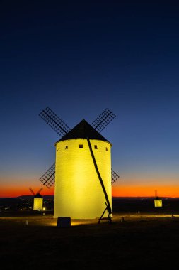 Yel değirmenleri tepede duruyor, alacakaranlıkta renkli bir günbatımı ufkuyla aydınlatılıyor.
