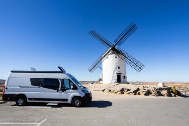 Beyaz karavan, açık mavi gökyüzünün altında geleneksel bir İspanyol yel değirmeninin yakınına park edilmiş.