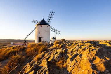 Beyaz yel değirmenleri İspanya 'nın Consuegra kentindeki kayalık bir tepede altın saat ışığıyla aydınlanıyor.