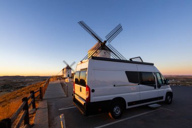 İspanya 'nın Consuegra kentindeki bir tepede, gün batımında tarihi yel değirmenlerinin yakınına park edilmiş bir karavan.