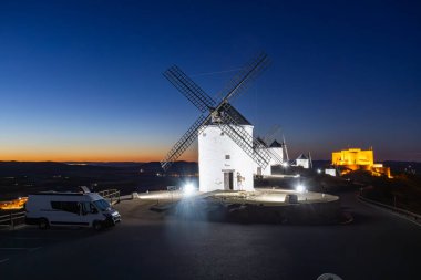 Geleneksel İspanyol yel değirmenleri ve mavi saatte bir tepede yakındaki bir karavanı aydınlatan bir şato.