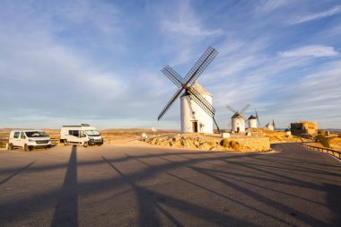 Günbatımında antik yel değirmenleri ve modern karavan araçlarının yer aldığı manzara.