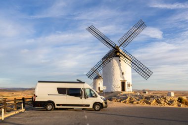 Kamp aracı mavi gökyüzünün altında ikonik İspanyol yel değirmenlerinin yanına park edilmiş. Özgürlüğü ve seyahati simgeliyor.