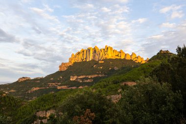 Montserrat sıradağlarının zirveleri bulutlu bir gökyüzünün altında altın ışıkla aydınlanır.