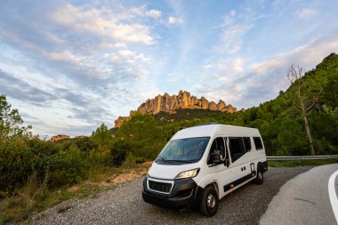 Arka planda Montserrat sıradağları olan dolambaçlı bir yolda park etmiş beyaz bir karavan.