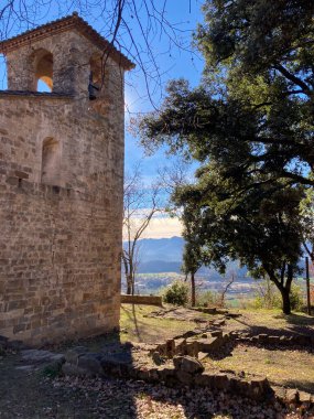 Antik taş çan kulesi, güneşli bir günde ağaçlarla ve harabelerle dolu bir vadi manzarasına bakıyor.