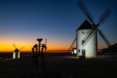 Don Kişot heykeli günbatımında geleneksel yel değirmenleriyle dramatik gökyüzüne karşı duruyor.