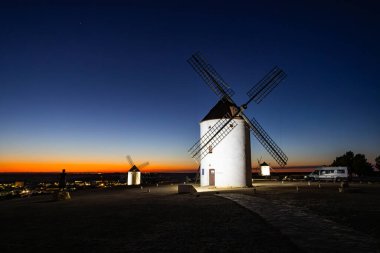 Tarihsel İspanyol yel değirmenleri bir tepede duruyor ve alacakaranlıkta Mota del Cuervo manzarasını seyrediyorlar.