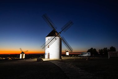 Tarihsel yel değirmenleri alacakaranlıkta canlı turuncu ve mavi gökyüzüyle tepede duruyor.