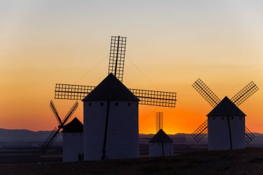Bir tepede duran yel değirmenleri canlı turuncu ve sarı günbatımına karşı siluetler oluşturuyor.