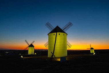 Geleneksel İspanyol yel değirmenleri, La Mancha 'da canlı bir günbatımına ve mavi saatli gökyüzüne karşı aydınlanıyor.