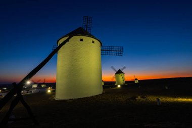 Yel değirmenleri gün batımında aydınlanan tepelerde duruyor, canlı bir alacakaranlık ufku yaratıyorlar.