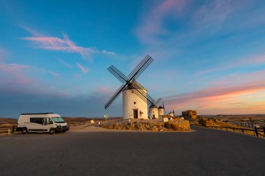 Renkli bir günbatımında, tepedeki tarihi yel değirmenlerinin yakınına park edilmiş bir karavan.