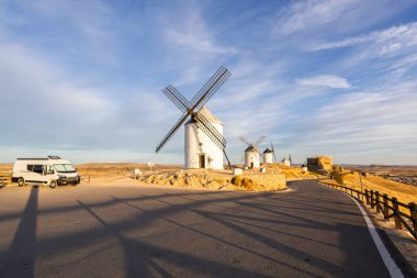 İspanya, Consuegra 'da günbatımında mavi gökyüzünün altında tarihi yel değirmenlerinin yakınına park edilmiş bir karavan.