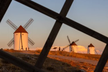 Mota del Cuervo, İspanya; 21 Eylül 2025: Mota del Cuervo yel değirmenleri günbatımı gökyüzünün altında altın renginde parlıyordu, ön plandaki ahşap çitlerle çerçevelenmişti.