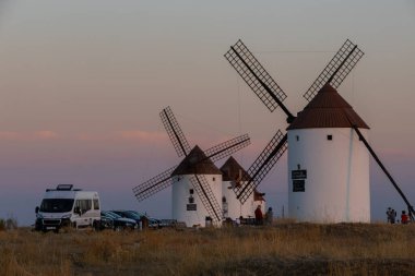 Mota del Cuervo, İspanya; 21 Eylül 2025: Pastel gökyüzünün altında park etmiş bir karavan ve ziyaretçilerle duran geleneksel İspanyol yel değirmenleri
