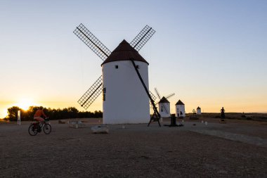 Mota del Cuervo, İspanya; 22 Eylül 2025: Bisikletçi huzurlu bir günbatımında tarihi İspanyol rüzgar değirmenlerinin önünden geçiyor