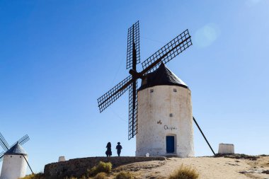 Consuegra, İspanya; 23 Eylül 2025: İnsanlar açık mavi gökyüzü altında geleneksel İspanyol yel değirmenleriyle kuru bir arazinin tadını çıkarıyorlar