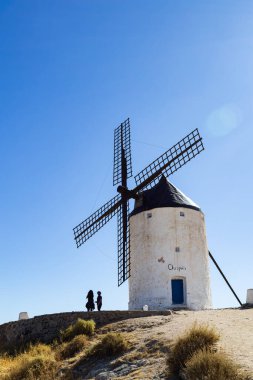 Consuegra, İspanya; 23 Eylül 2025: İnsanlar Consuegra 'da açık mavi bir gökyüzünün altında geleneksel beyaz bir yel değirmeni görüyorlar