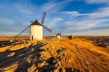 Consuegra, İspanya; 24 Eylül 2025: Consuegra yel değirmenleri gün batımında mavi gökyüzü ve yumuşak bulutlarla altın bir tepenin üzerinde duruyor